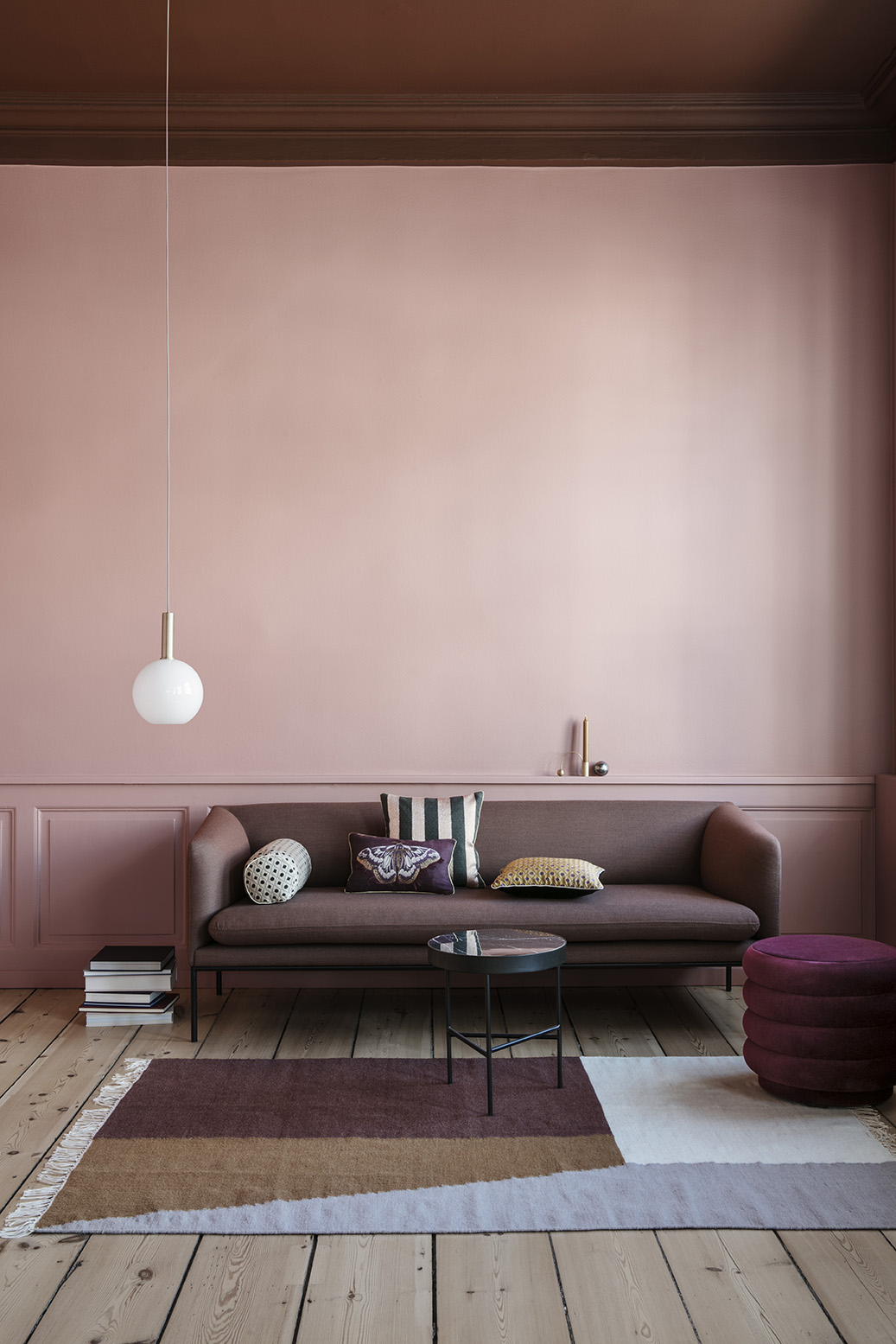 Støvet lyserød stue med høje paneler i samme farve. Mørk bordeaux sofa står op af væggen med mønstrede puder i.