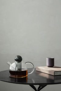 På billedet ses en tekande i glas ra Ferm Living placeret samme men en kop på et bord foran en støvet grøn væg