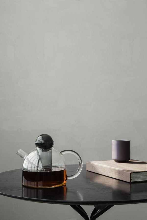 På billedet ses en tekande i glas ra Ferm Living placeret samme men en kop på et bord foran en støvet grøn væg
